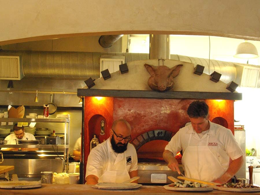 Two chefs make pizzas in front of a wood burning oven