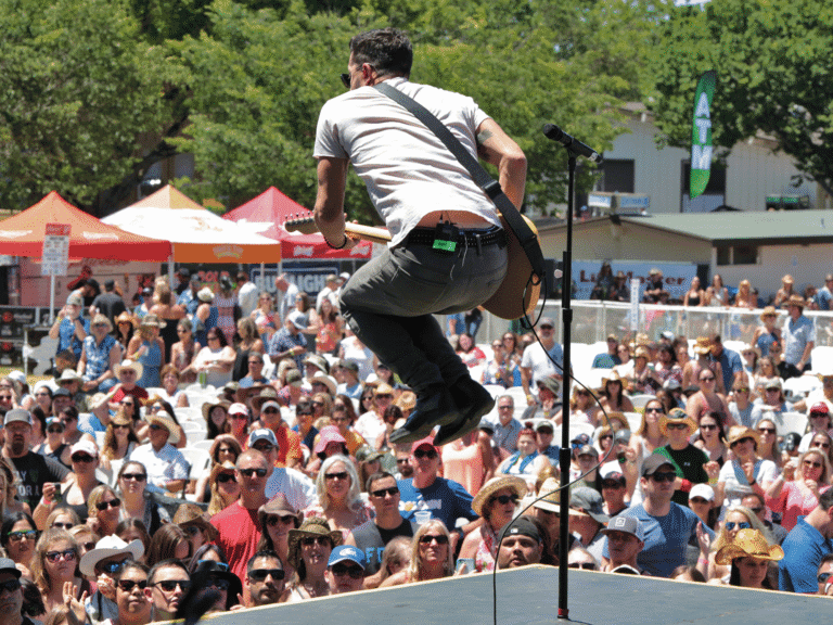 musician jumping with guitar while playing on stage in front of large crowd at country summer music festival