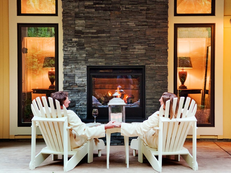 A couple in white bathrobes sit in white deck chairs in front of an outdoor fireplace set in a yellow facade at Farmhouse Inn and Spa in Sonoma County, California