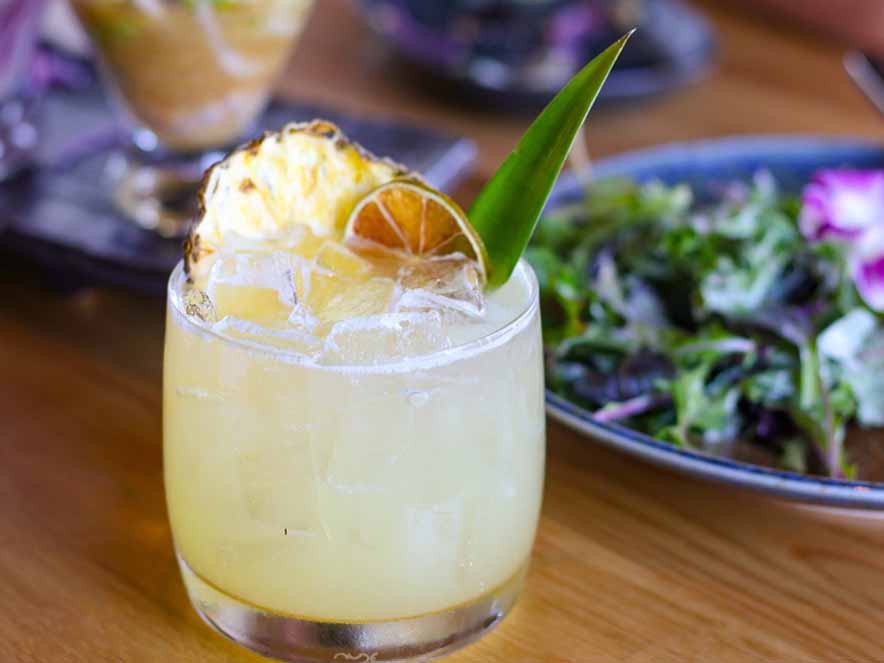 yellow drink with pineapple and orange in glass with food in the background