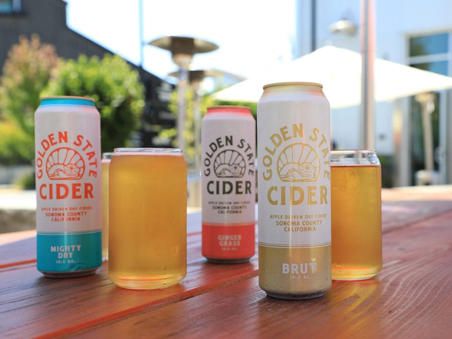 Cans of cider and full glasses sit on a table in the sun