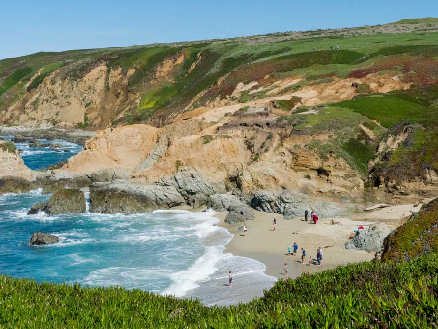 People stand on a beach in a cove surrounded by big, green hills