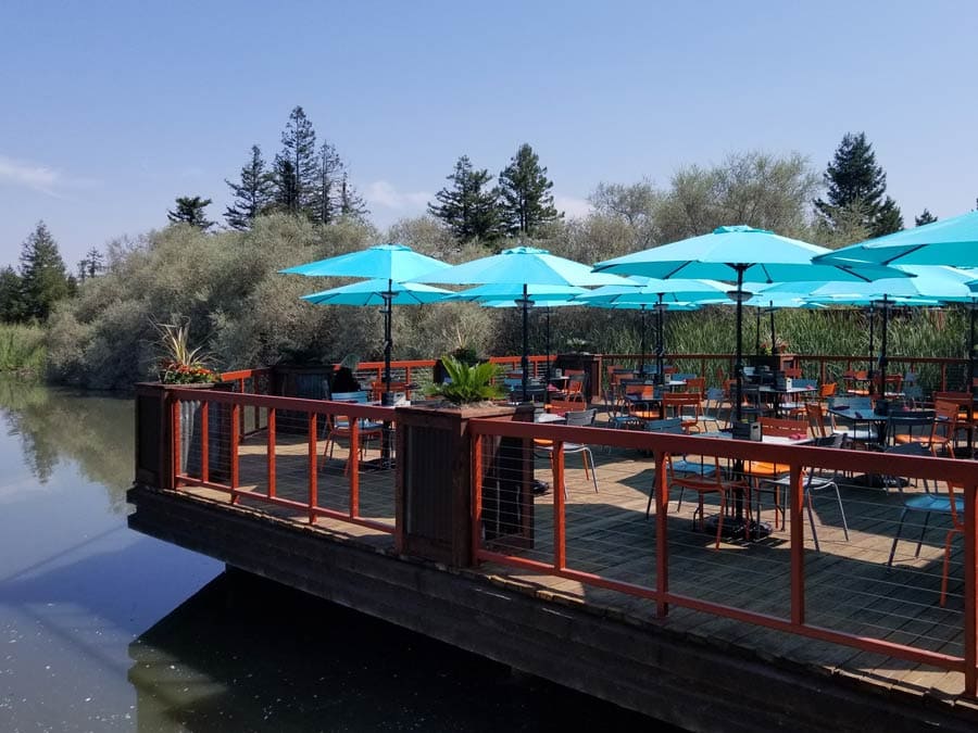 Tables with blue umbrellas are set on the sunny deck overlooking the lake in Sonoma County