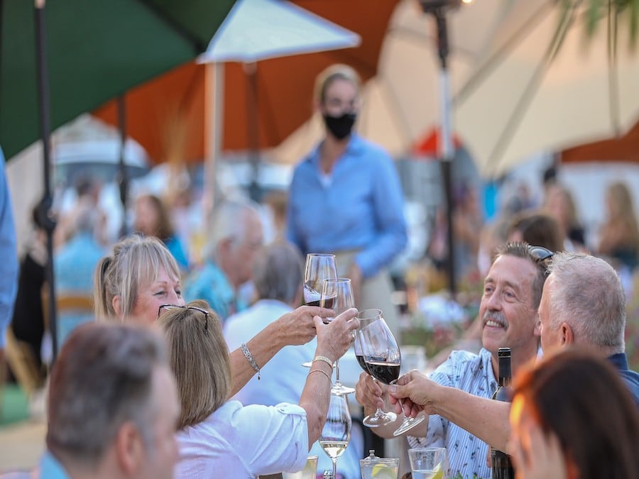 People at an outdoor table toasting each other with full wine glasses while a waitress in a mask remains out of focus in the background