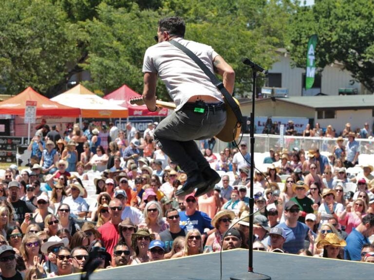 Guy jumping with guitar while playing on stage in front of large crowd