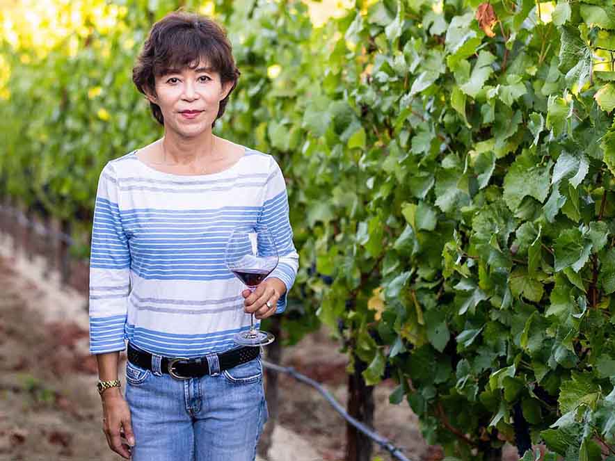 Picture of Asian American woman standing beside vineyards holding wine glass